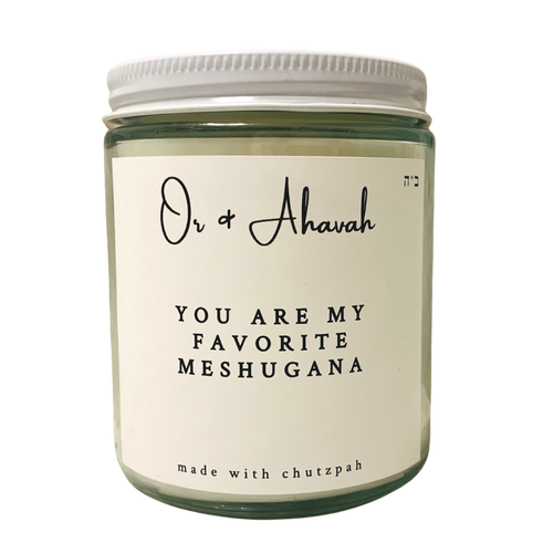 Clear glass candle with a white metal lid. Brand name is Or & Ahavah. Candle name is You Are My Favorite Meshugana. At the bottom, it says made with chutzpah. Scent notes are sea salt, cardamom seed, amber, and clove.