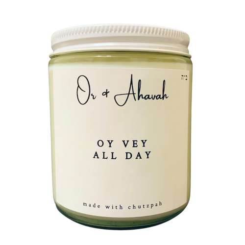 Clear glass candle with a white metal lid. Brand name is Or & Ahavah. Candle name is Oy Vey All Day. At the bottom, it says made with chutzpah. Scent notes are lemon peel, lemon verbena, and lemongrass.