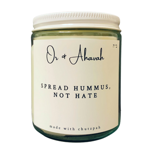 Clear glass candle with a white metal lid. Brand name is Or & Ahavah. Candle name is Spread Hummus, Not Hate. At the bottom, it says made with chutzpah. Scent notes are sandalwood, patchouli, honey, and amber.