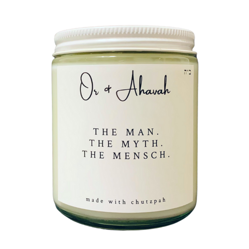 Clear glass candle with a white metal lid. Brand name is Or & Ahavah. Candle name is The Man, The Myth, The Mensch. At the bottom, it says made with chutzpah. Scent notes are oakmoss, sage, tonka bean, and amber.