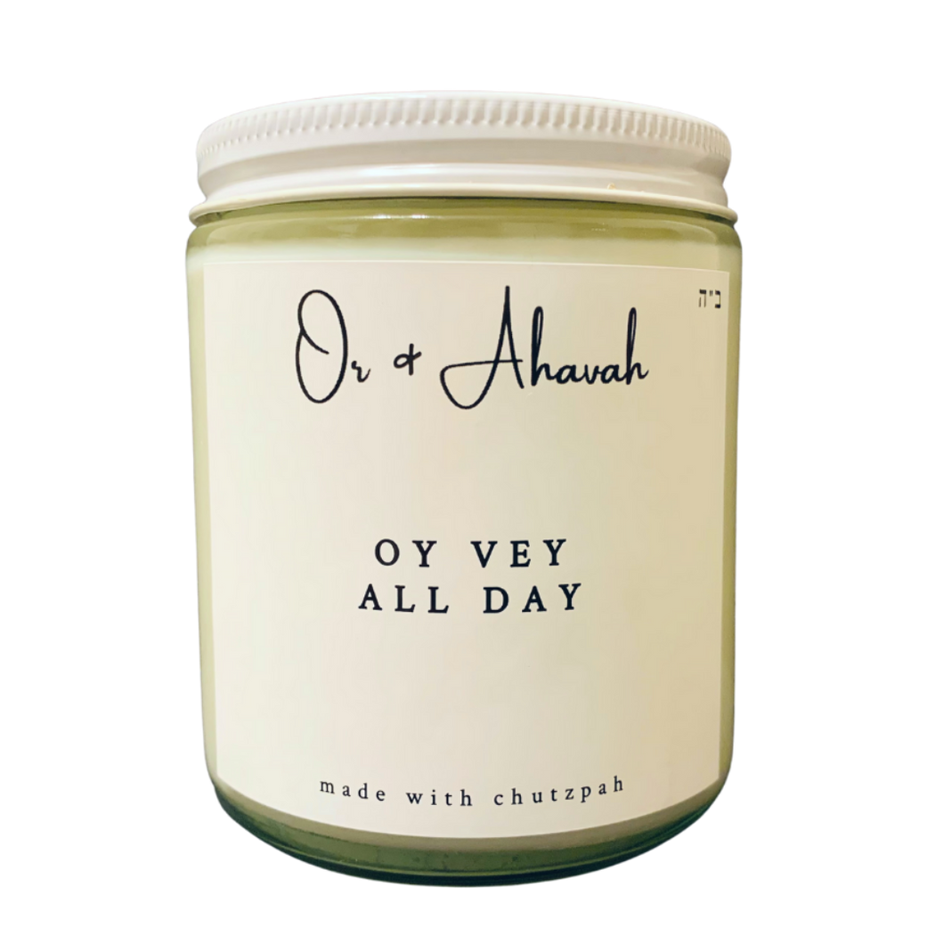 Clear glass candle with a white metal lid. Brand name is Or & Ahavah. Candle name is Oy Vey All Day. At the bottom, it says made with chutzpah. Scent notes are lemon peel, lemon verbena, and lemongrass.